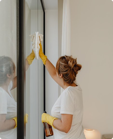 Kobieta w żółtych rękawiczkach myje dużą szybę i trzyma butelkę z płynem do czyszczenia szyb.