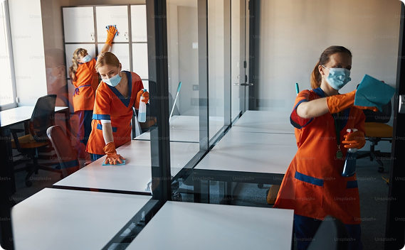 Trzech pracowników w maseczkach i pomarańczowych uniformach czyści biuro. Sprzątają biurka i szklane ścianki działowe, używając ściereczek i płynów.