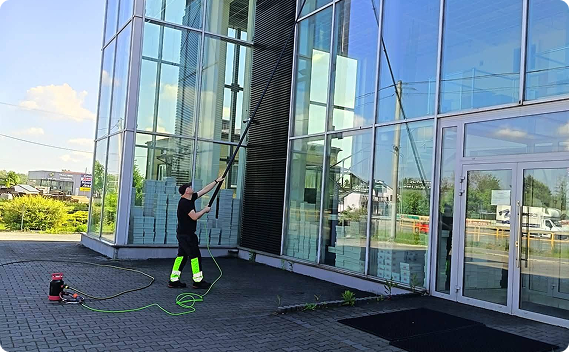 Pracownik w profesjonalnym stroju roboczym (z odblaskowymi elementami) myje wysoką szklaną elewację nowoczesnego budynku, używając specjalistycznego kija teleskopowego.